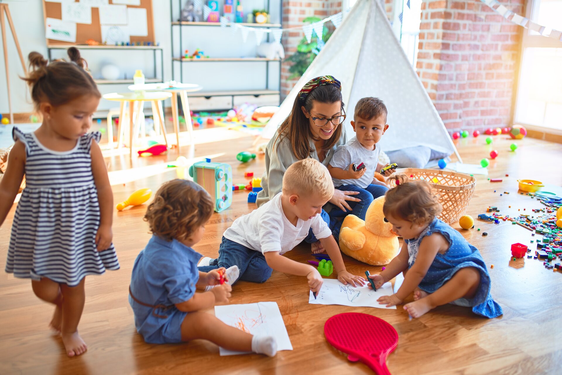 Lehrerin und Gruppe von Kleinkindern im Kindergarten, die auf dem Boden laufen, sitzen und mit Papier und Bleistift zeichnen.